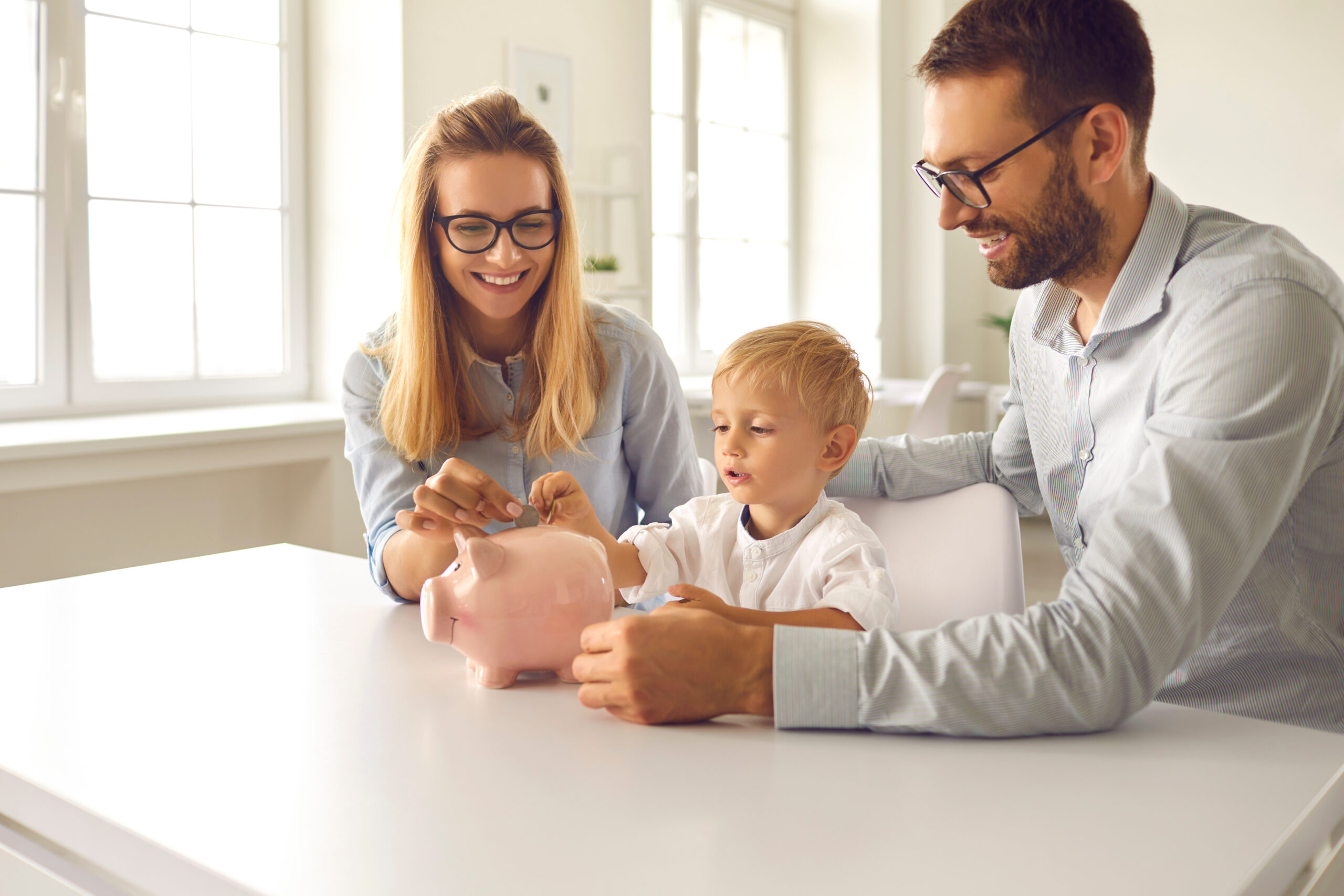 Financial literacy. Little boy with young parents puts coins in a piggy bank sitting at a table in the room. Mom and dad teach their son to effectively manage the budget and spend money wisely.