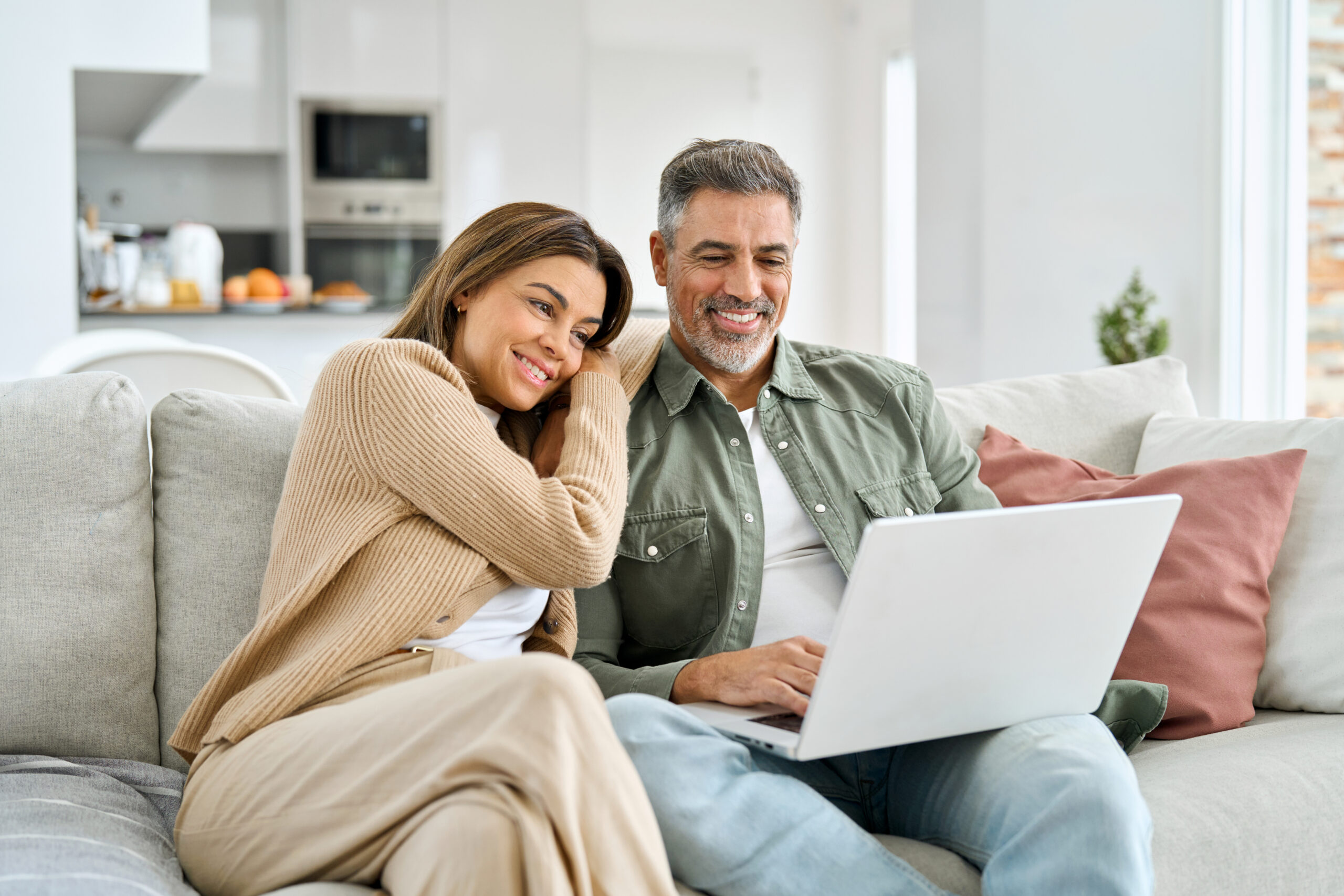 Happy middle aged couple using laptop relaxing on couch at home. Smiling mature man and woman looking at computer watching video, browsing or shopping in ecommerce store sitting on sofa in living room